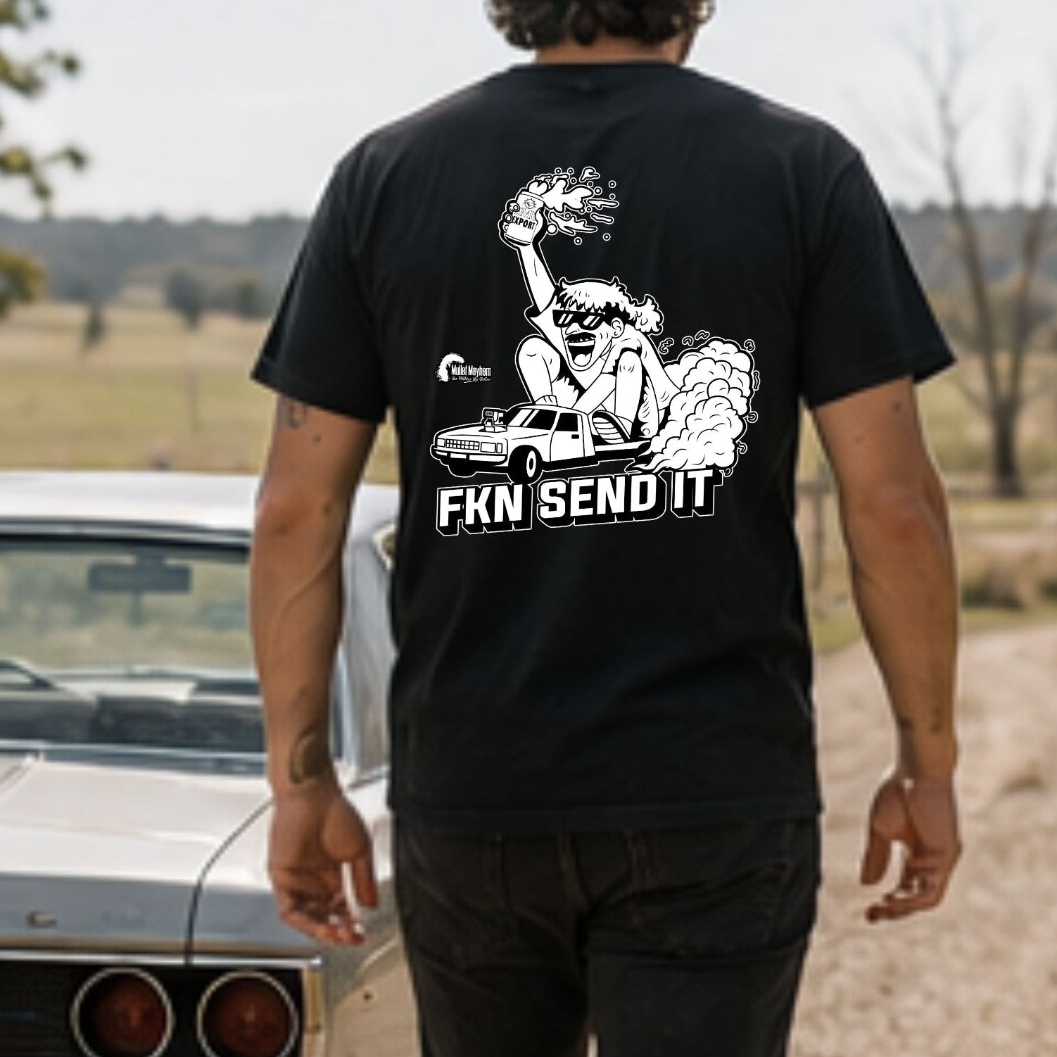 A man viewed from behind walking on a dirt road wearing a black t-shirt with a large white graphic print on the back that reads 'FKN SEND IT!'. He is standing next to a classic silver car in an outdoor rural setting.