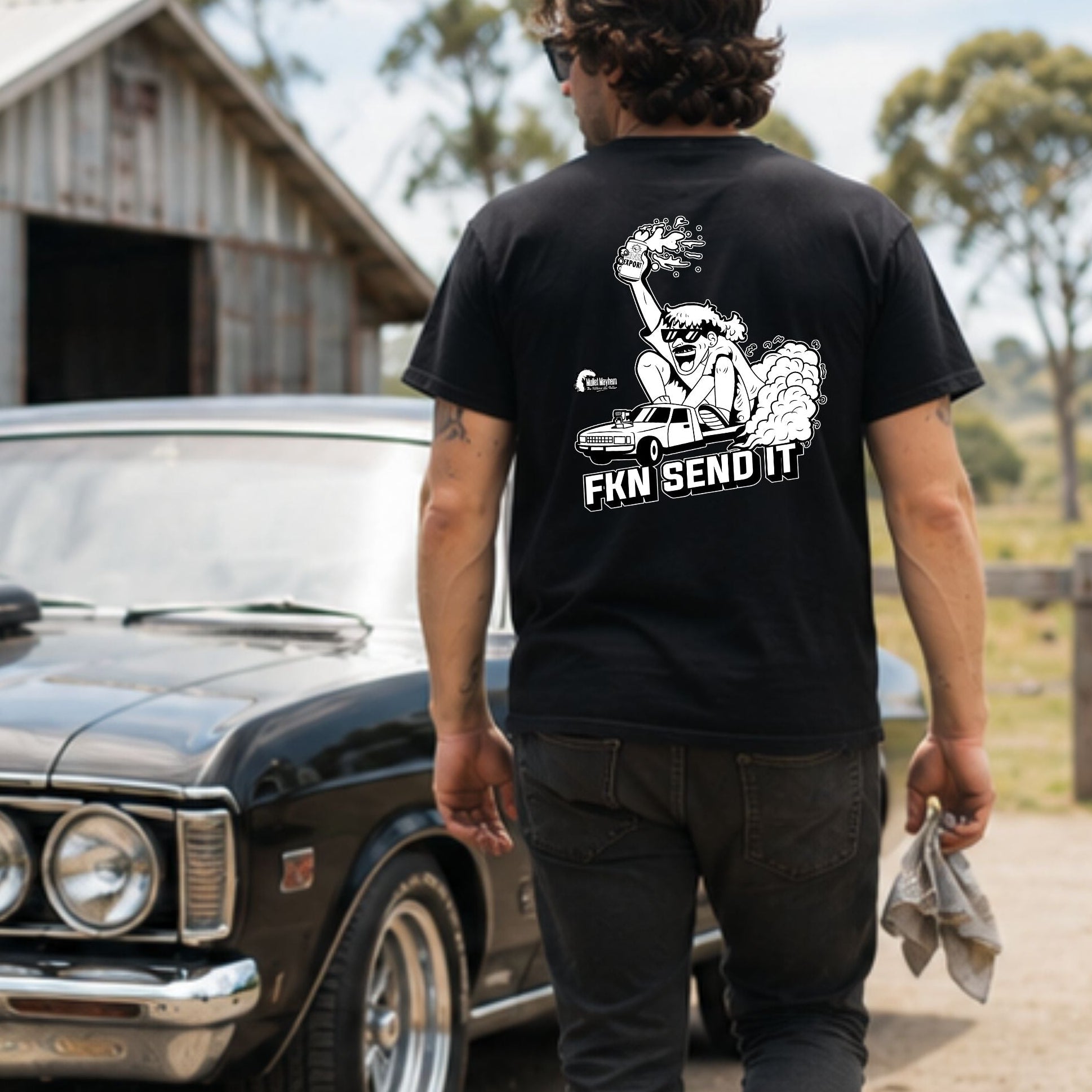 A man wearing a black graphic t-shirt with a white fist design and the text 'FKN SEND IT' stands in front of a black classic car in an outdoor rural setting with a barn in the background.