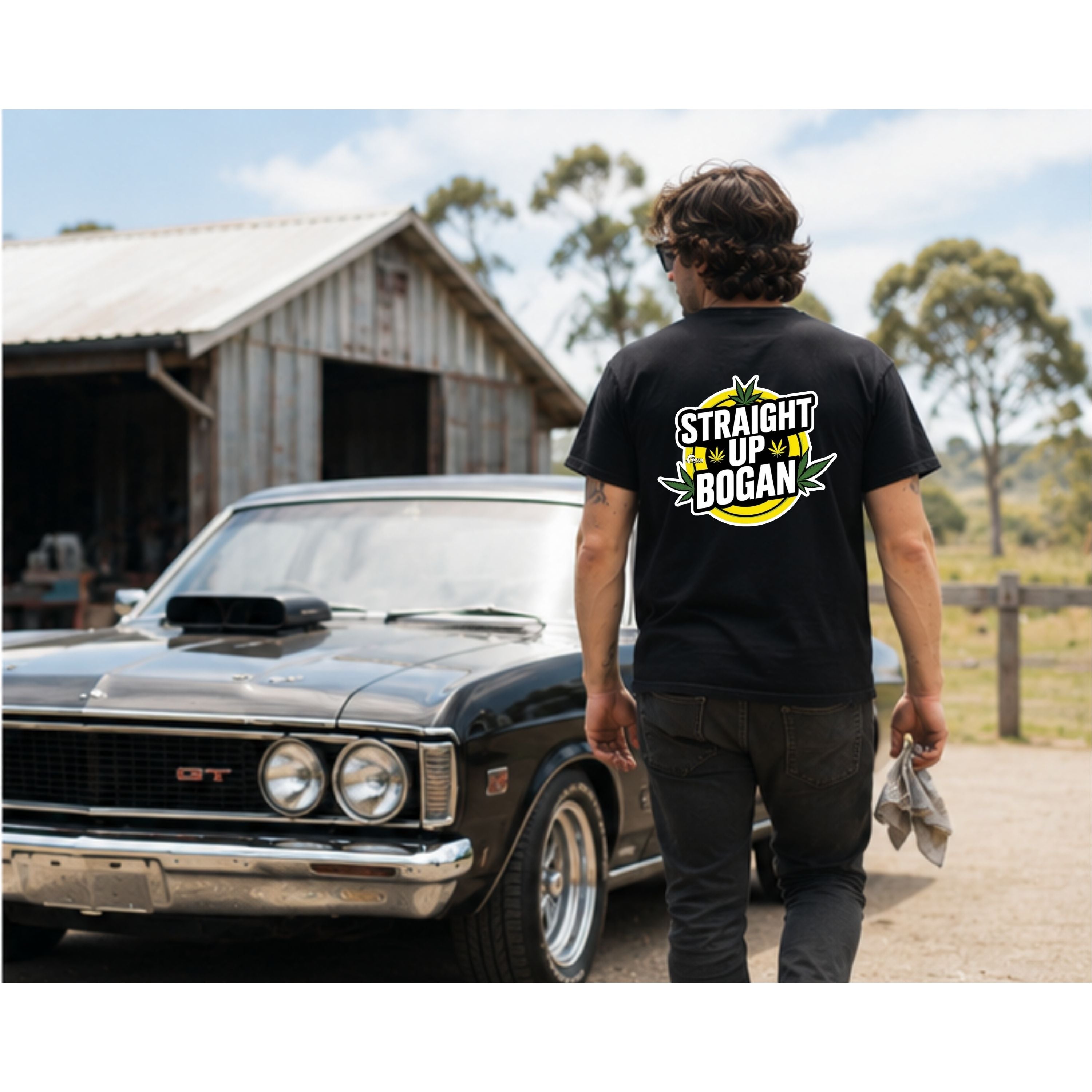 A man wearing a black graphic t-shirt with a 'STRAIGHT UP BOGAN' design on the back, standing next to a classic black car in front of a rustic barn.