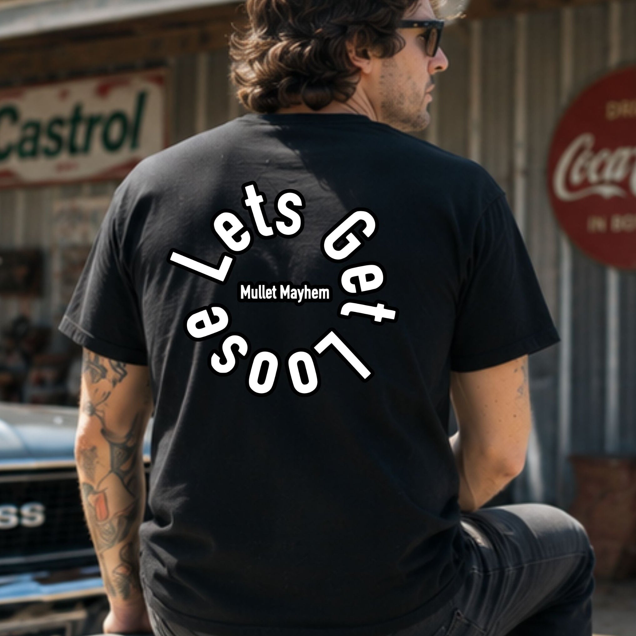 A man wearing a black t-shirt with a white graphic text design on the back, sitting in front of a rustic metal building with vintage Coca-Cola and Castrol signs.