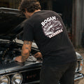 A man wearing a black t-shirt with a white 'BOGAN SKIDDERS' graphic on the back, leaning over the open hood of a classic car in a garage setting.