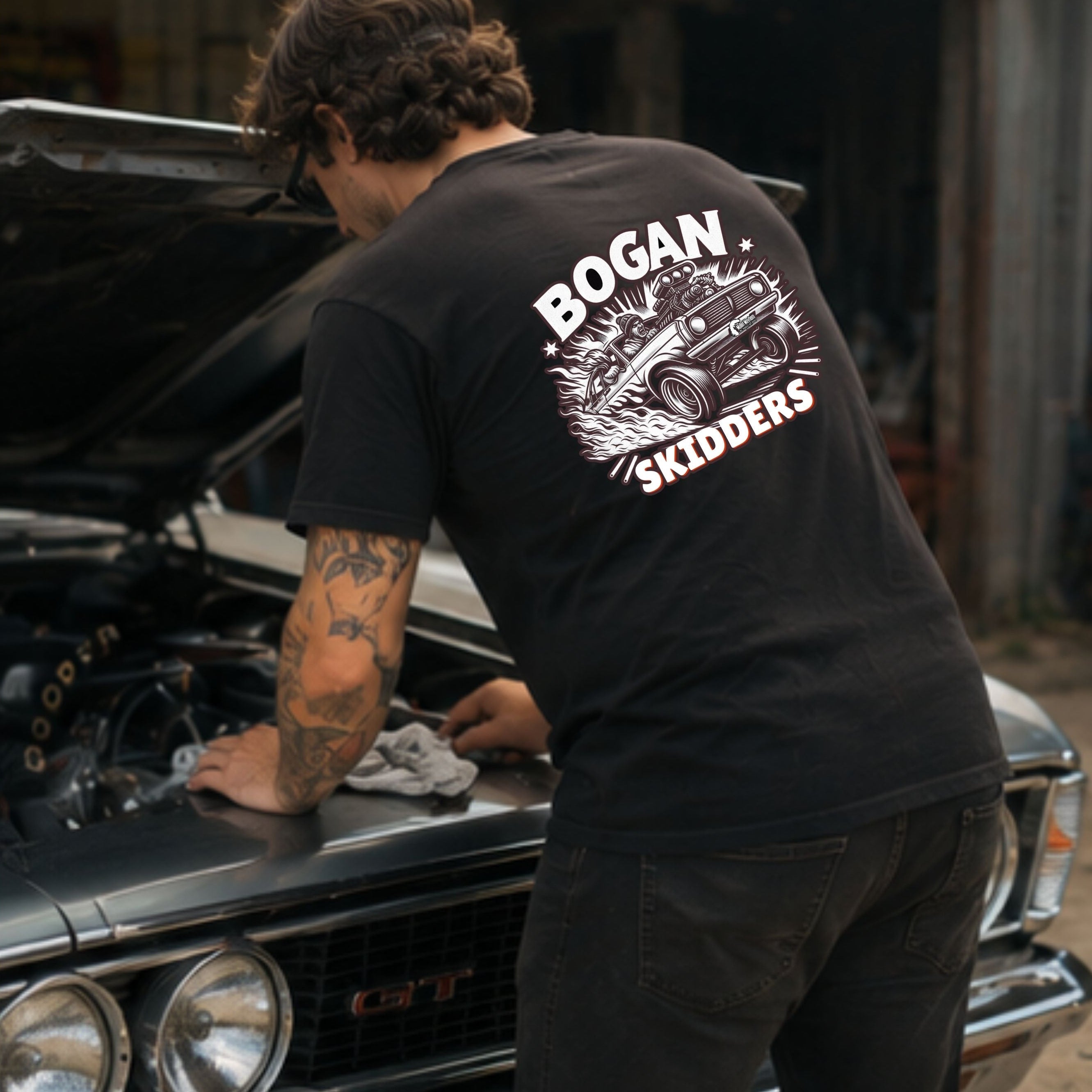 A man wearing a black t-shirt with a white 'BOGAN SKIDDERS' graphic on the back, leaning over the open hood of a classic car in a garage setting.