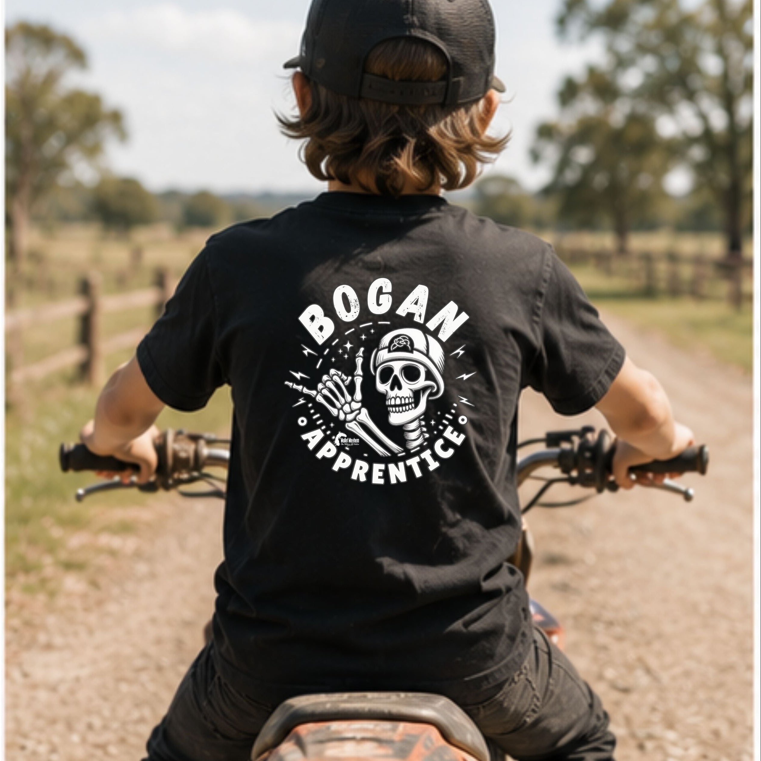 A child seen from behind wearing a black t-shirt with a white 'BOGAN APPRENTICE' skull graphic, sitting on a dirt bike on a country path.