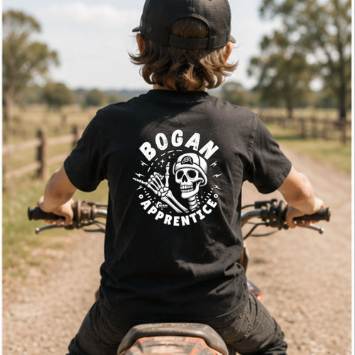 A child seen from behind wearing a black t-shirt with a white 'BOGAN APPRENTICE' skull graphic, sitting on a dirt bike on a country path.
