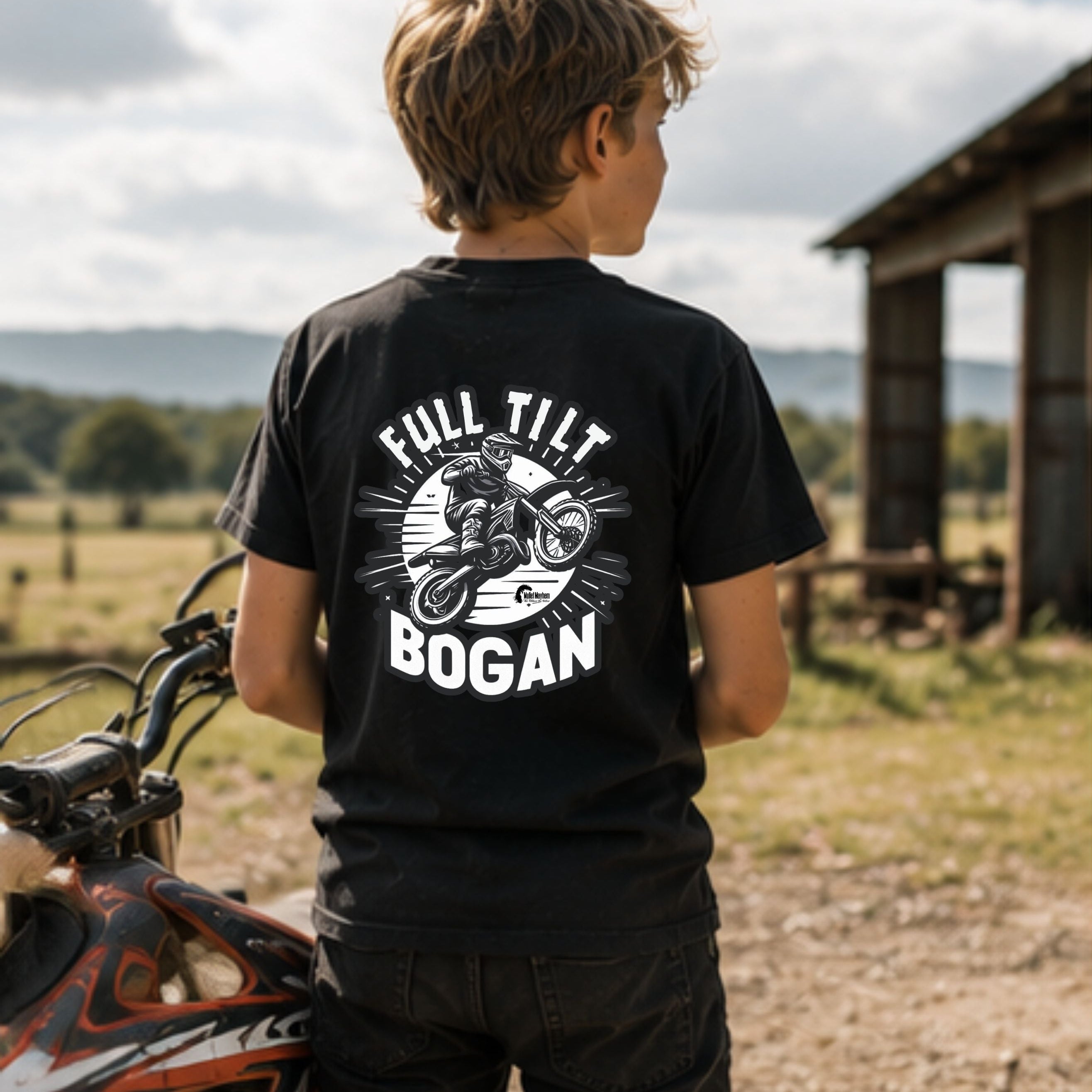 A boy seen from behind wearing a black graphic t-shirt with a white 'FULL TILT BOGAN' motorcycle print, standing outdoors next to a dirt bike in a rural setting.