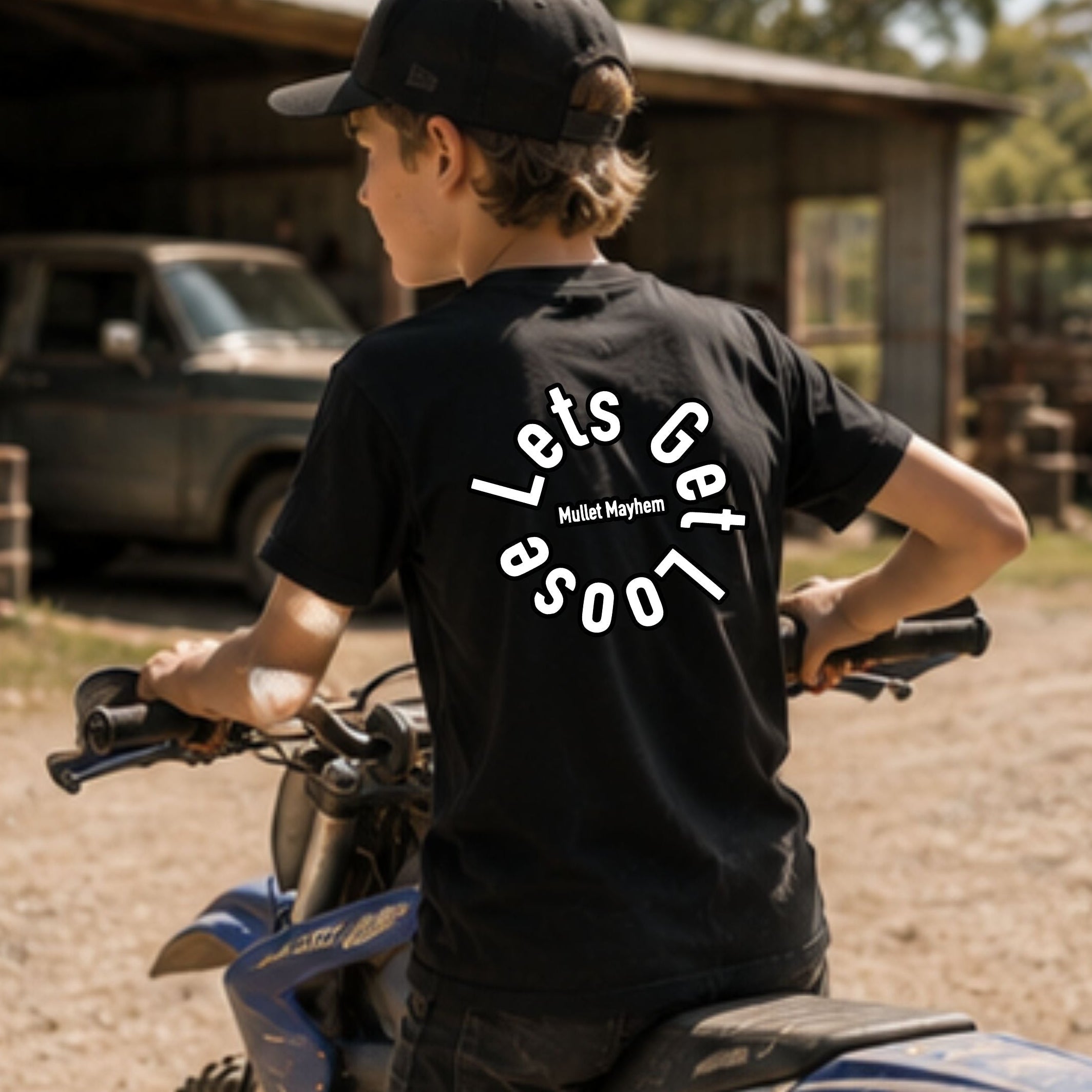 A boy wearing a black t-shirt with a white graphic on the back and a black cap, sitting on a blue dirt bike in an outdoor rural setting with a barn and truck in the background.