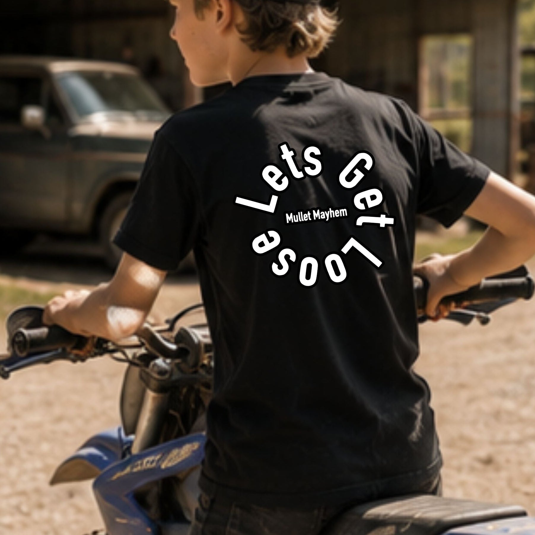 A boy wearing a black t-shirt with a white graphic on the back and a black cap, sitting on a blue dirt bike in an outdoor rural setting with a barn and truck in the background.