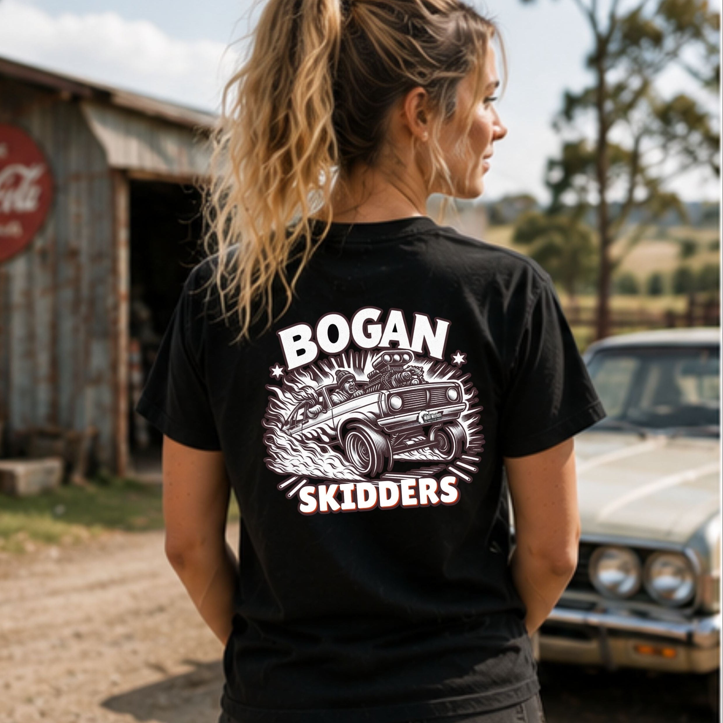 A woman wearing a black graphic t-shirt with a white and pink 'BOGAN SKIDDERS' print on the back, standing outdoors in front of a rustic barn and a vintage car.