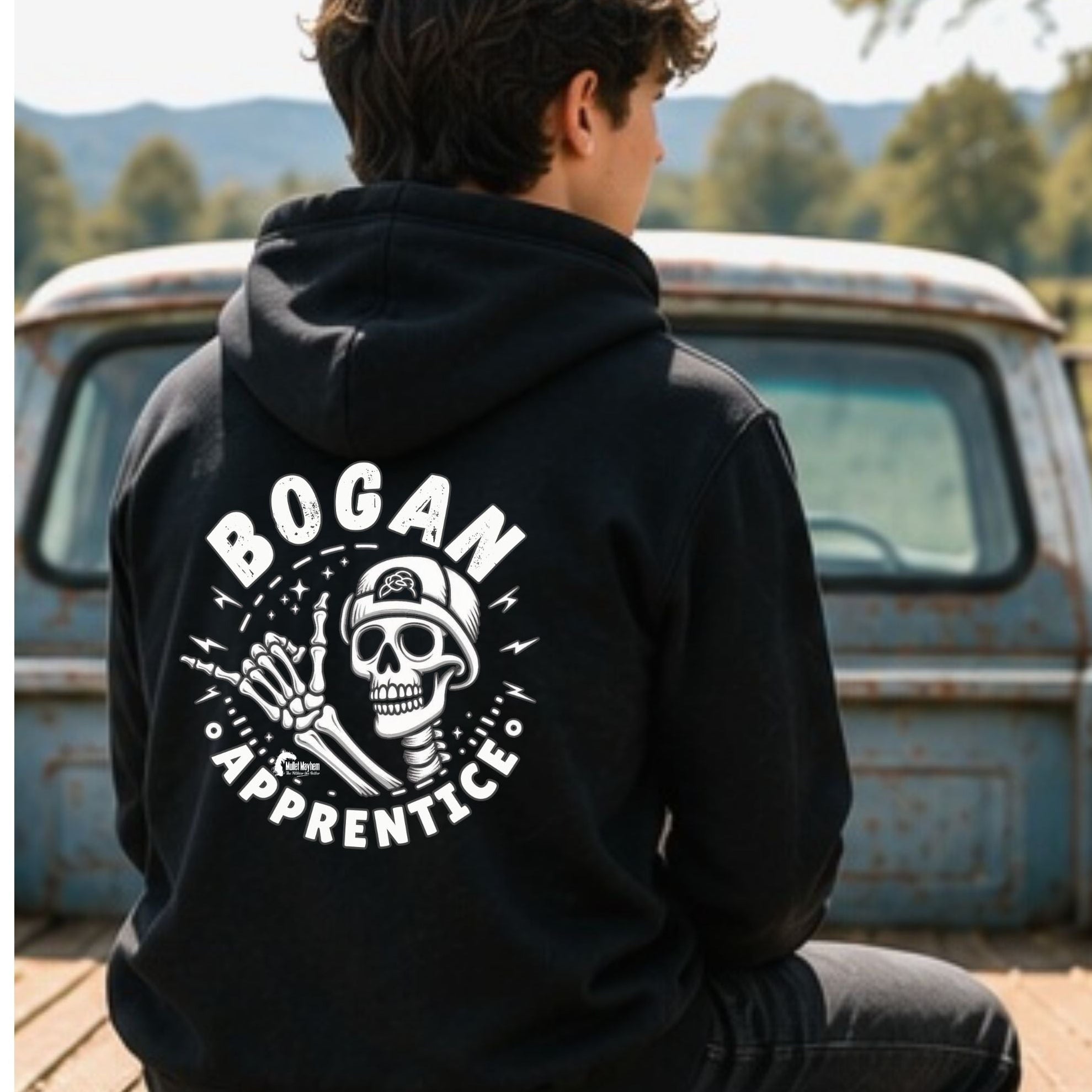 A person sitting on the tailgate of a vintage blue pickup truck, viewed from behind, wearing a black hoodie with a large white graphic on the back that reads 'BOGAN APPRENTICE' around a skull and crossed tools design.