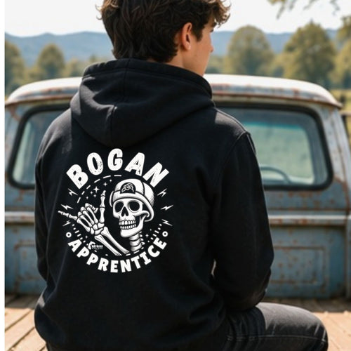 A person sitting on the tailgate of a vintage blue pickup truck, viewed from behind, wearing a black hoodie with a large white graphic on the back that reads 'BOGAN APPRENTICE' around a skull and crossed tools design.