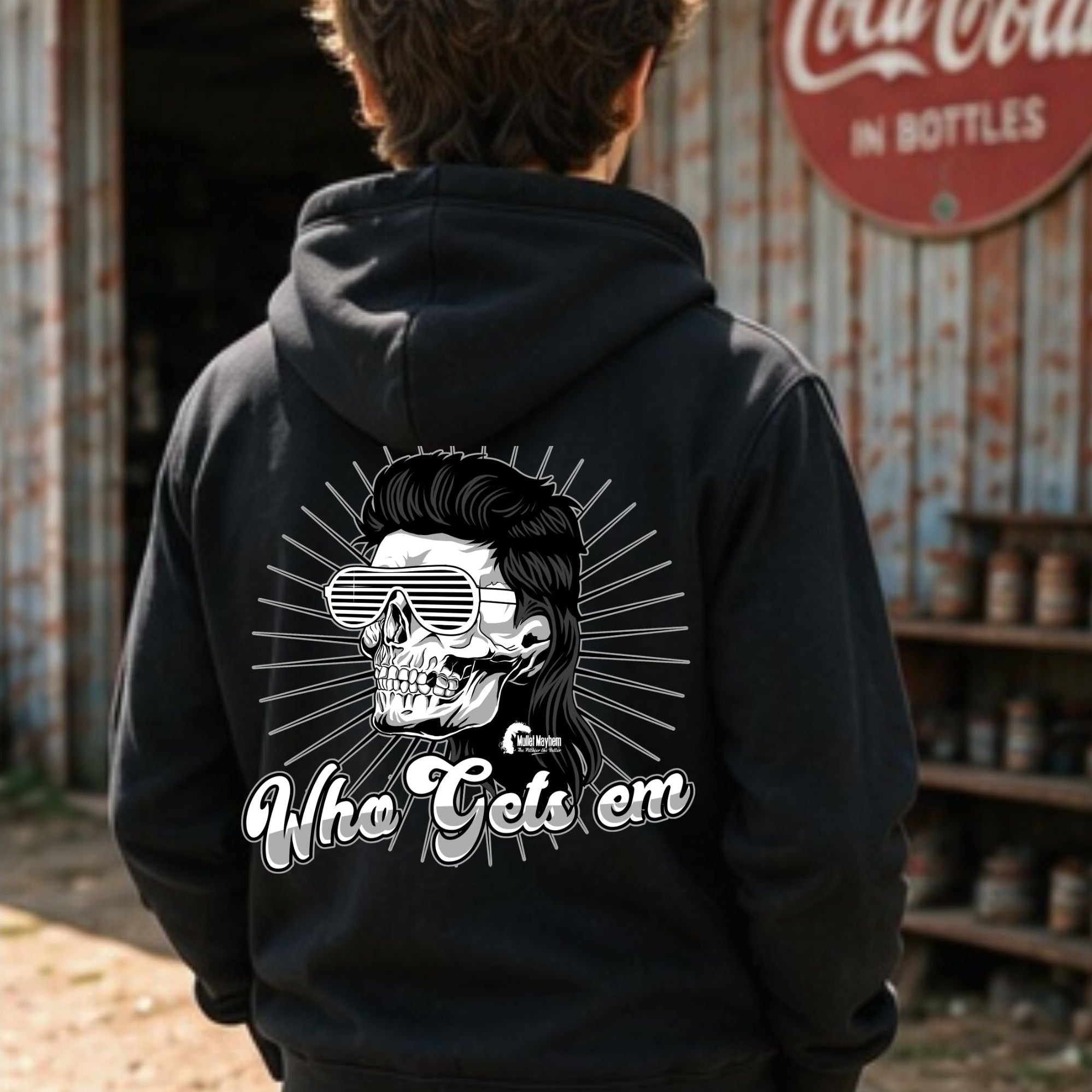 A person viewed from behind wearing a black hoodie with a large white graphic on the back featuring a skull in sunglasses and the text 'Who Gets em'. The background shows a rustic outdoor setting with a vintage Coca-Cola sign on a corrugated metal wall.