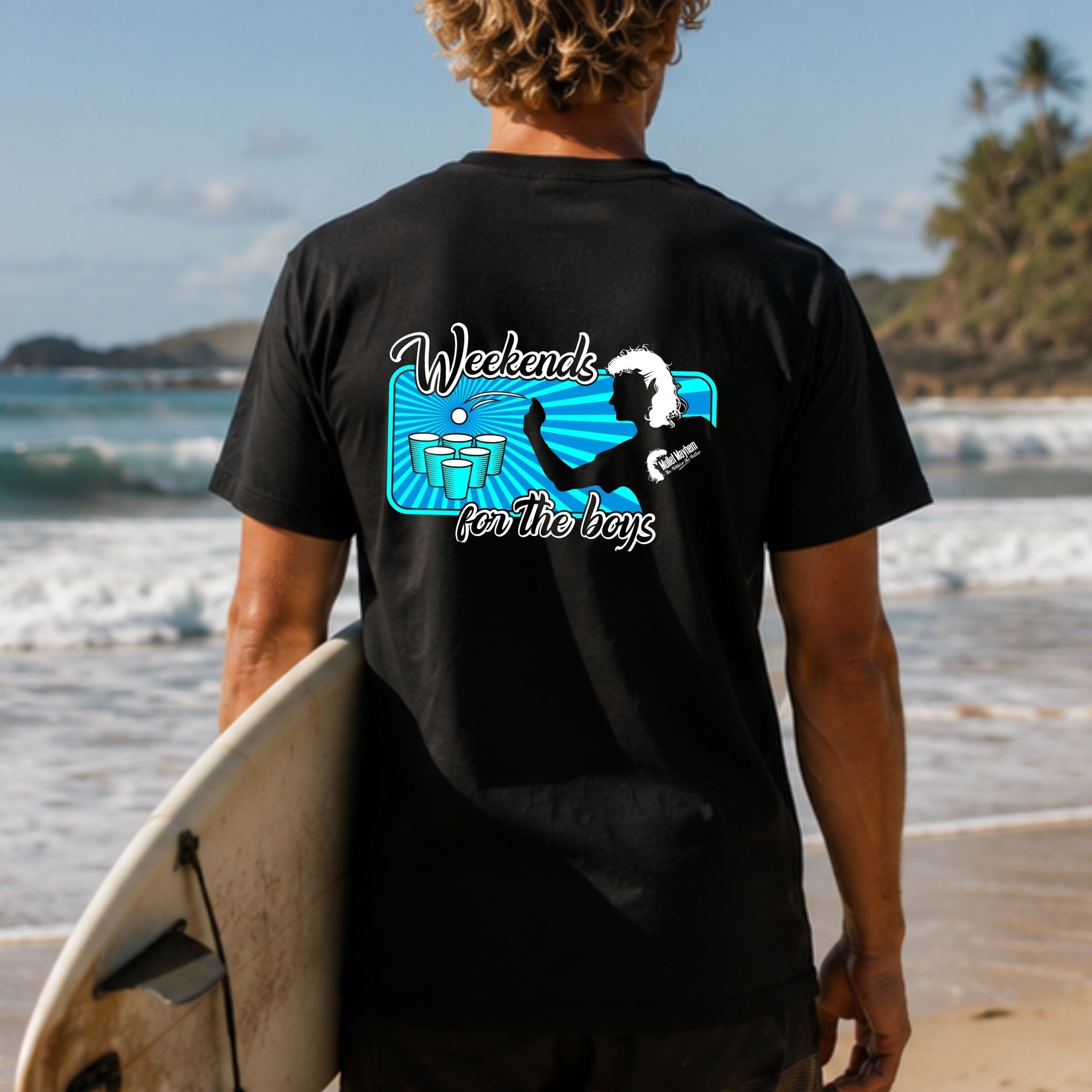 A man standing on a beach, viewed from behind, wearing a black t-shirt with a blue and white graphic on the back that reads 'Weekend for the boys' and holding a surfboard.