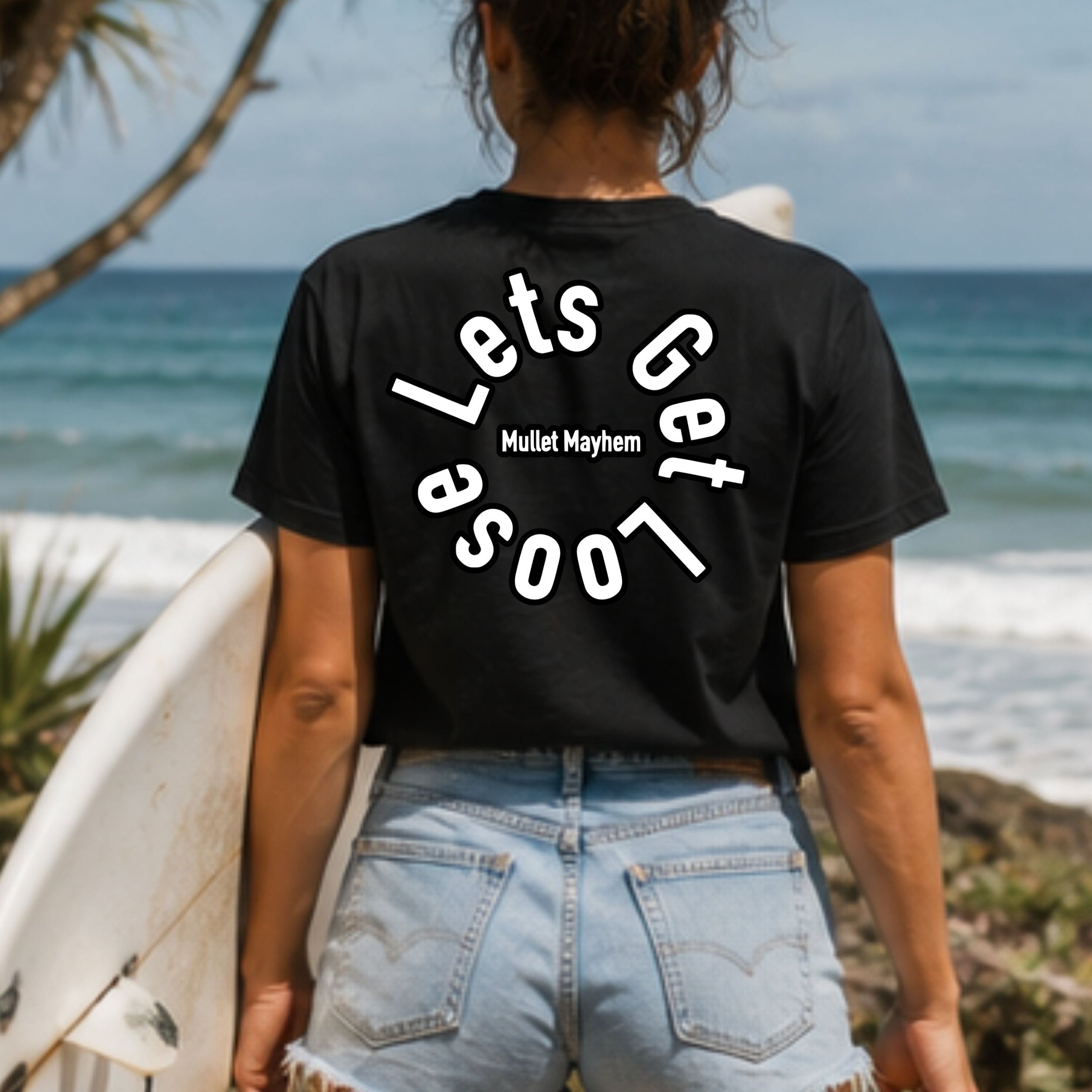 A woman standing on a beach with her back to the camera, wearing a black graphic t-shirt with 'Lets Get Loose' printed in white on the back. She is holding a white surfboard and wearing light-wash denim shorts.