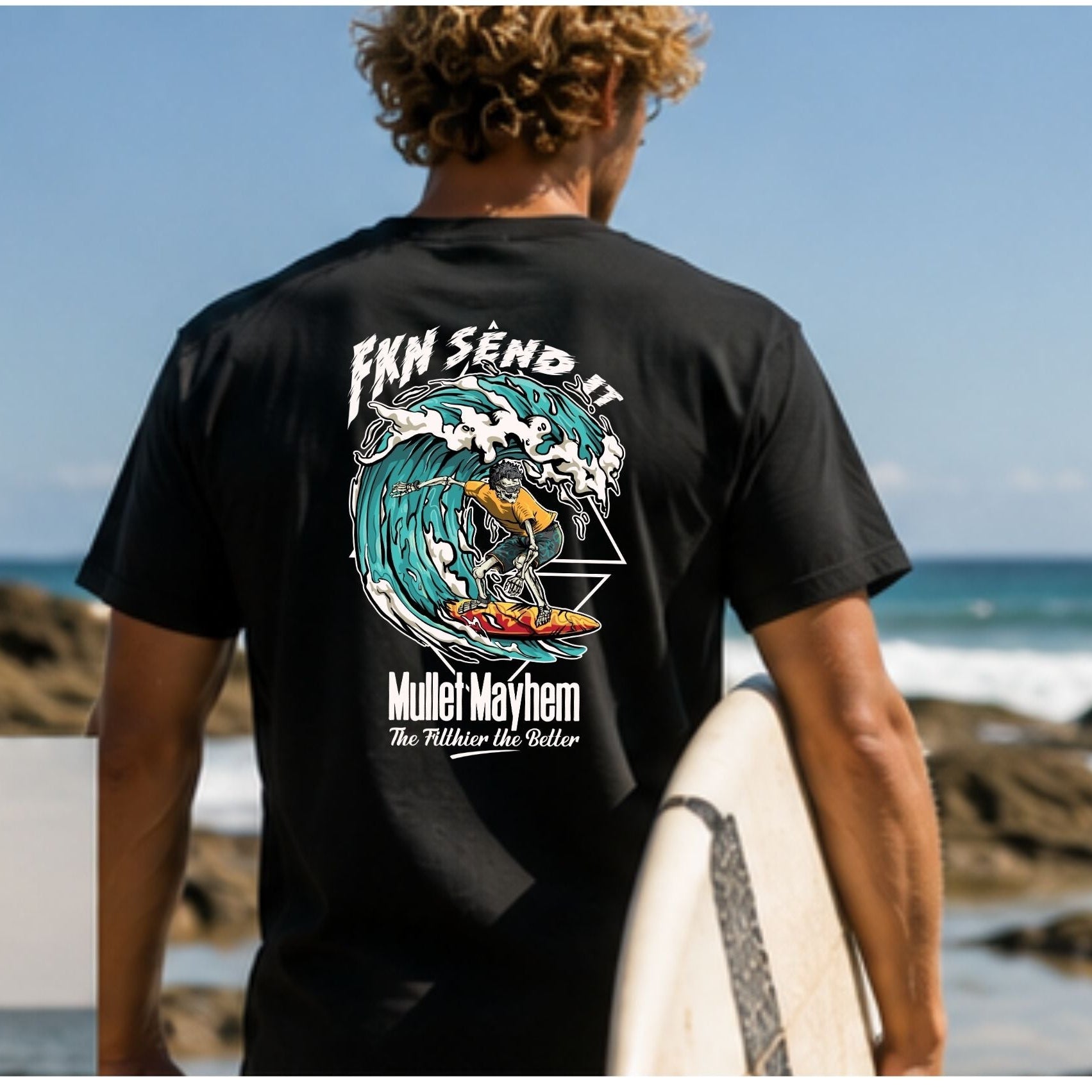 A man viewed from behind wearing a black t-shirt with a large graphic print on the back, standing on a rocky beach holding a surfboard. The graphic features a blue and white wave with the text 'FKK SEND IT' and 'Muler Mayhem'.