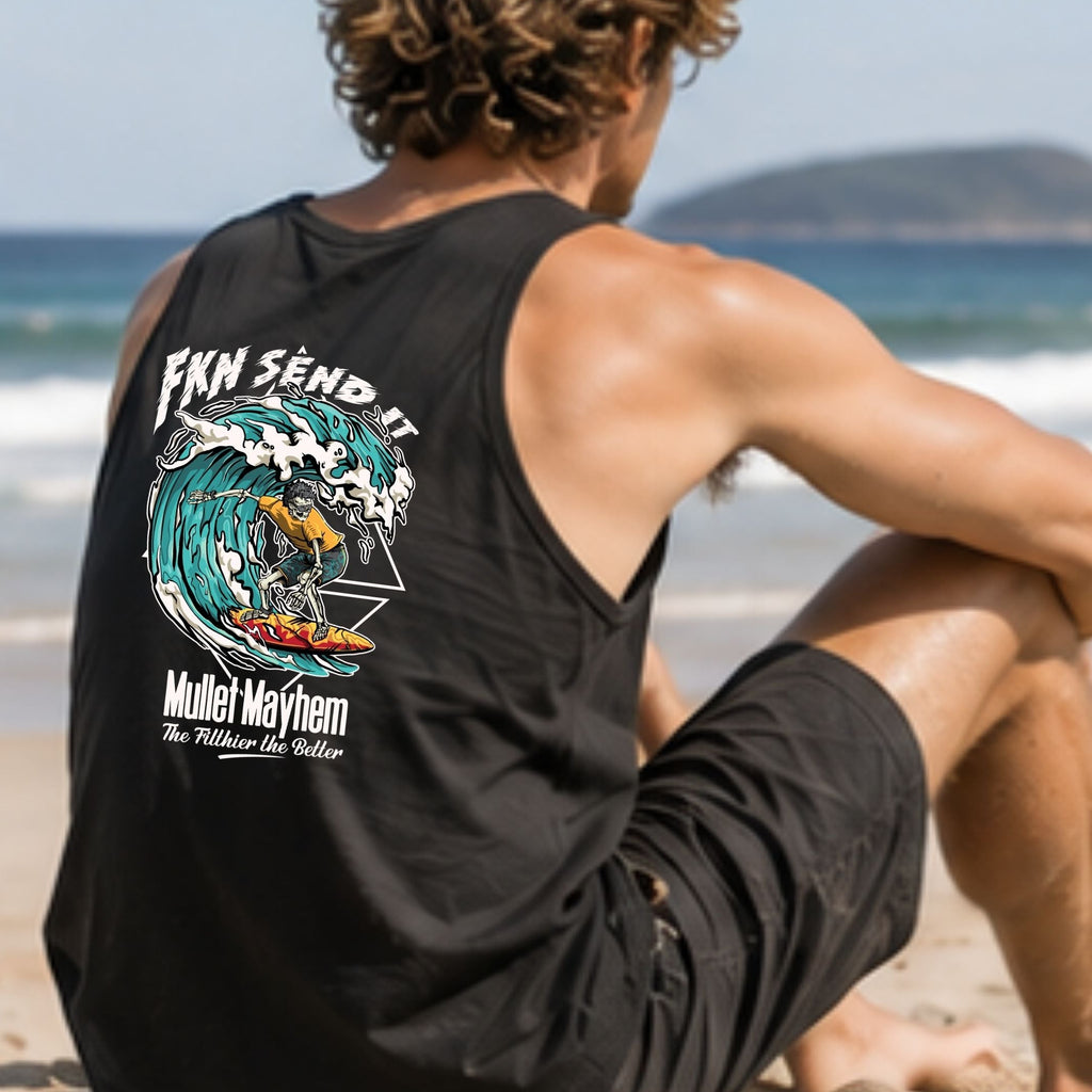 A man sits on a sandy beach facing the ocean, wearing a black tank top with a large multicolor wave graphic and text on the back, along with black shorts.