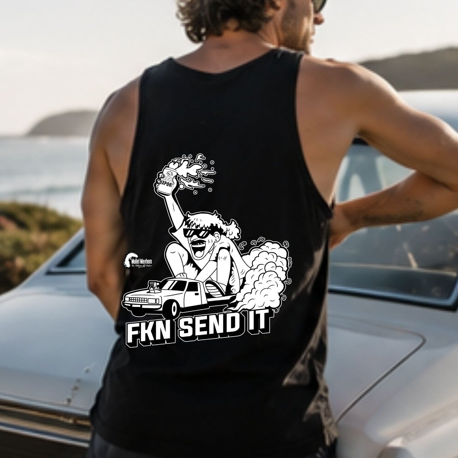 A man wearing a black tank top with a white graphic and the text 'FKN SEND IT' stands next to a vintage white car outdoors near the ocean.