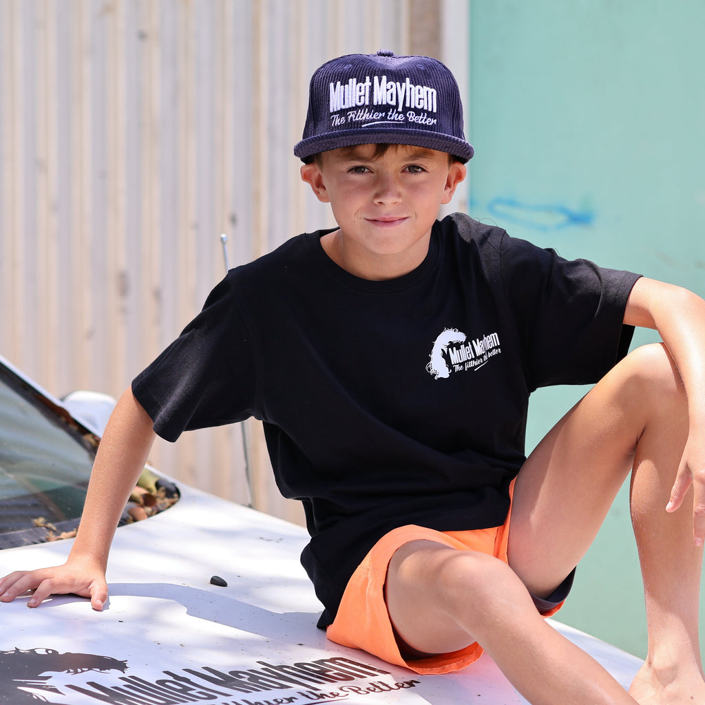 A young boy wearing a black t-shirt with a white logo, orange shorts, and a navy blue cap with 'Maiden Mayhem' printed on it, sitting on the hood of a white car.