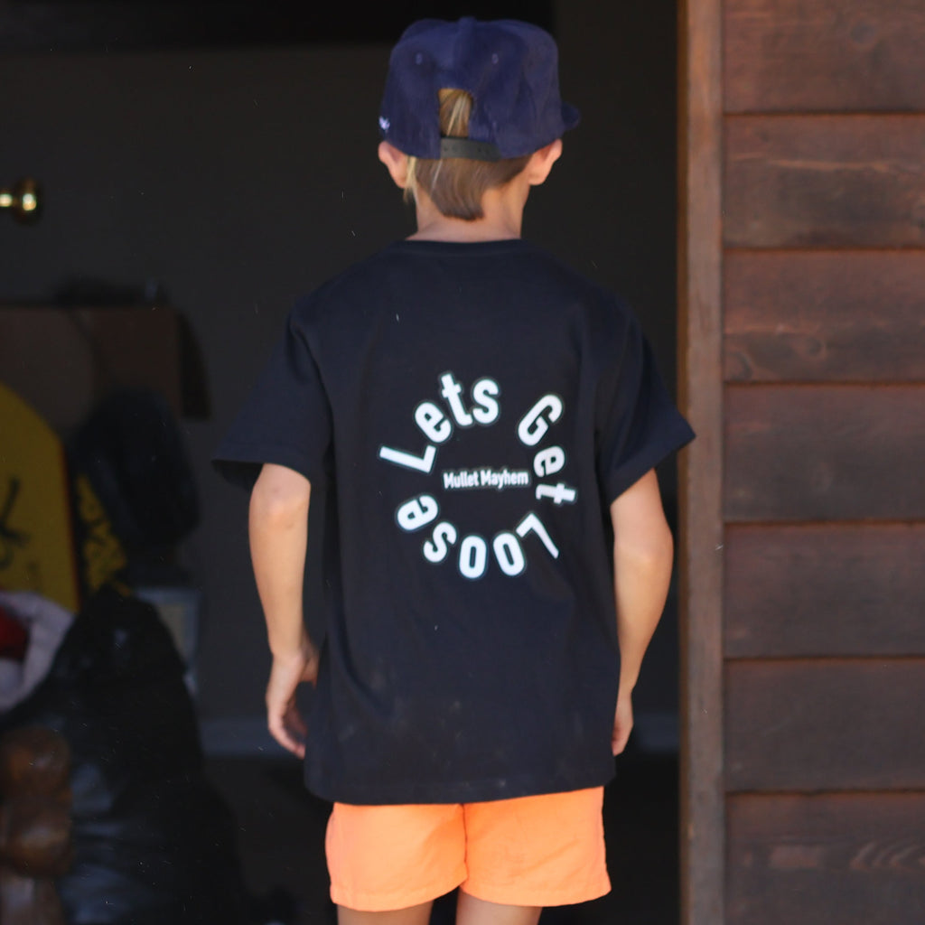 A person wearing a black graphic t-shirt with 'Loose Cigs' printed in white on the back, orange shorts, and a dark blue cap, standing in front of an open doorway of a rustic wooden building. A white dog is visible in the bottom right corner.