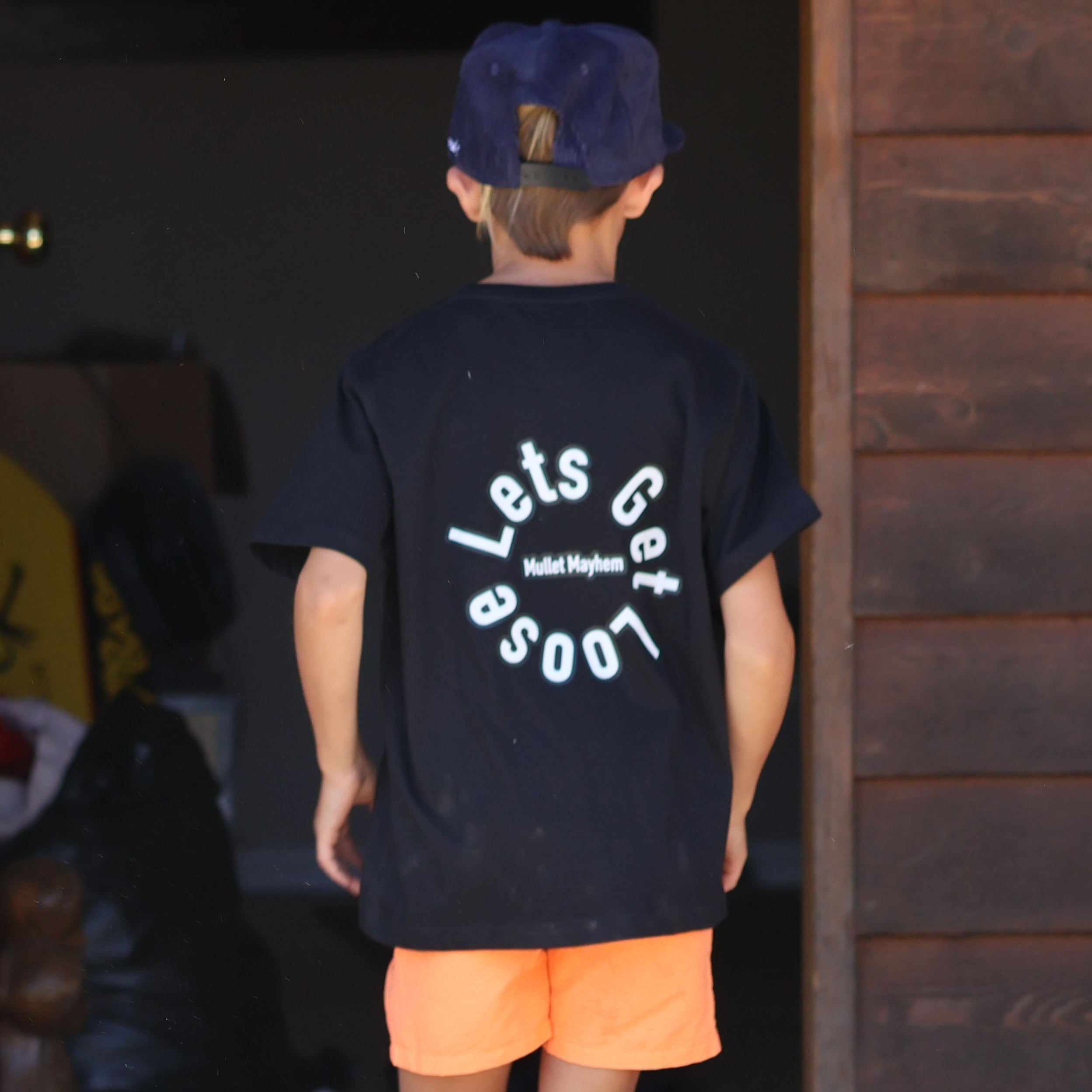 A person wearing a black graphic t-shirt with 'Loose Cigs' printed in white on the back, orange shorts, and a dark blue cap, standing in front of an open doorway of a rustic wooden building. A white dog is visible in the bottom right corner.