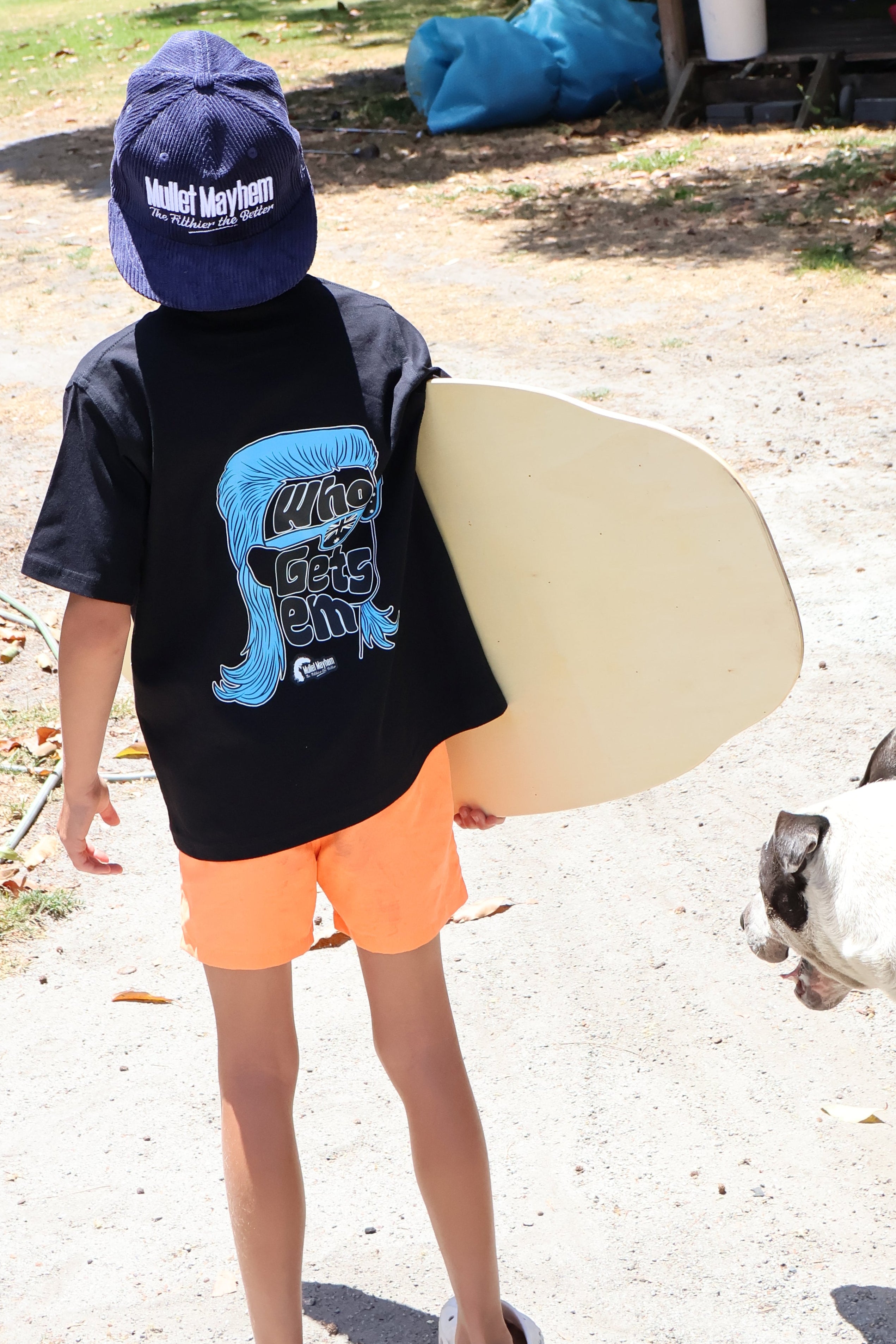 A child standing outdoors on a dirt path holding a light-colored surfboard, wearing a navy blue t-shirt with a blue graphic, orange shorts, and a navy blue cap. A white and brown dog is visible on the right side, and a small shelter with blue bags is in the background.
