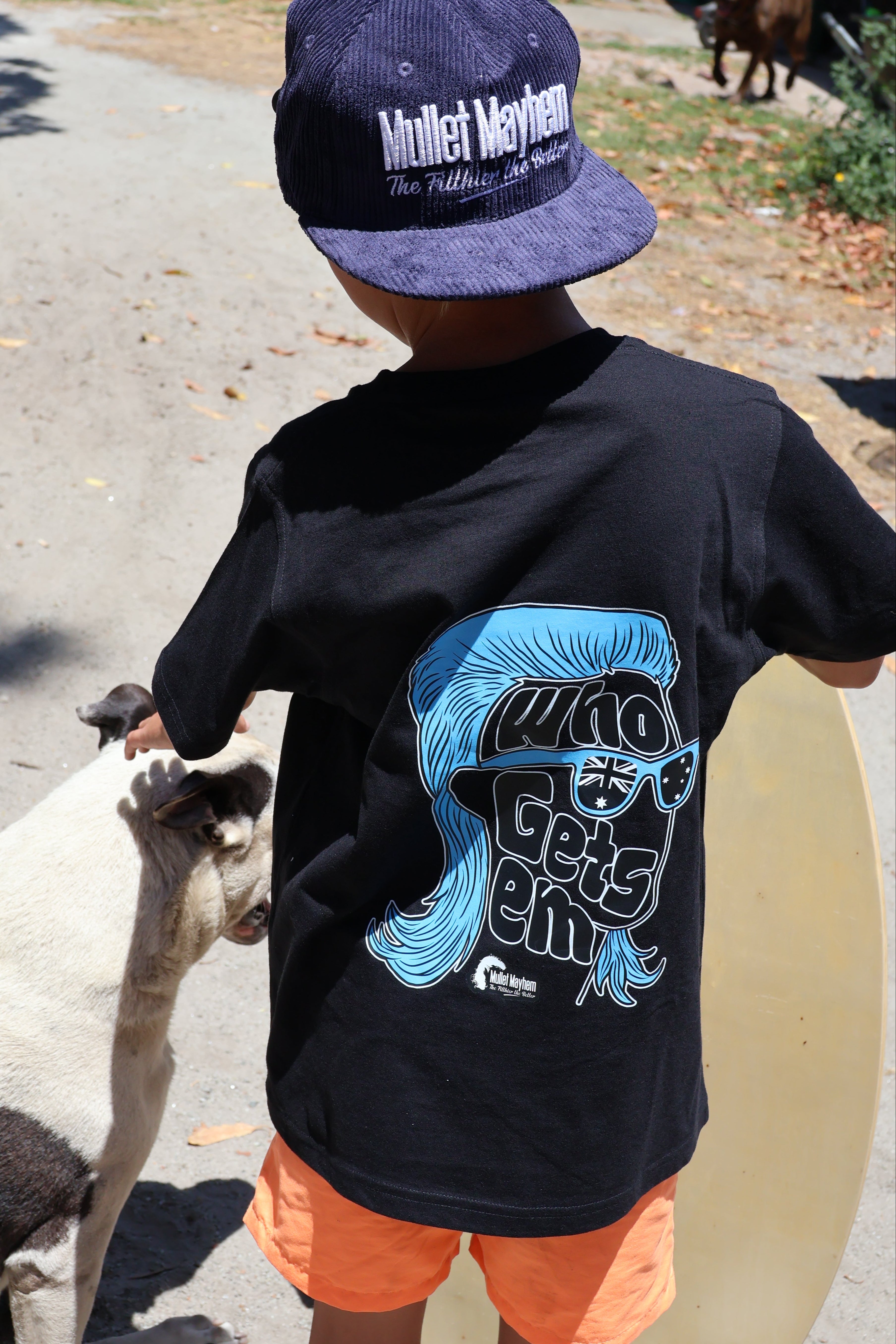 A child seen from behind wearing a black t-shirt with a blue and white graphic print, orange shorts, and a navy blue corduroy cap. The child is standing outdoors on a paved path next to a goat.