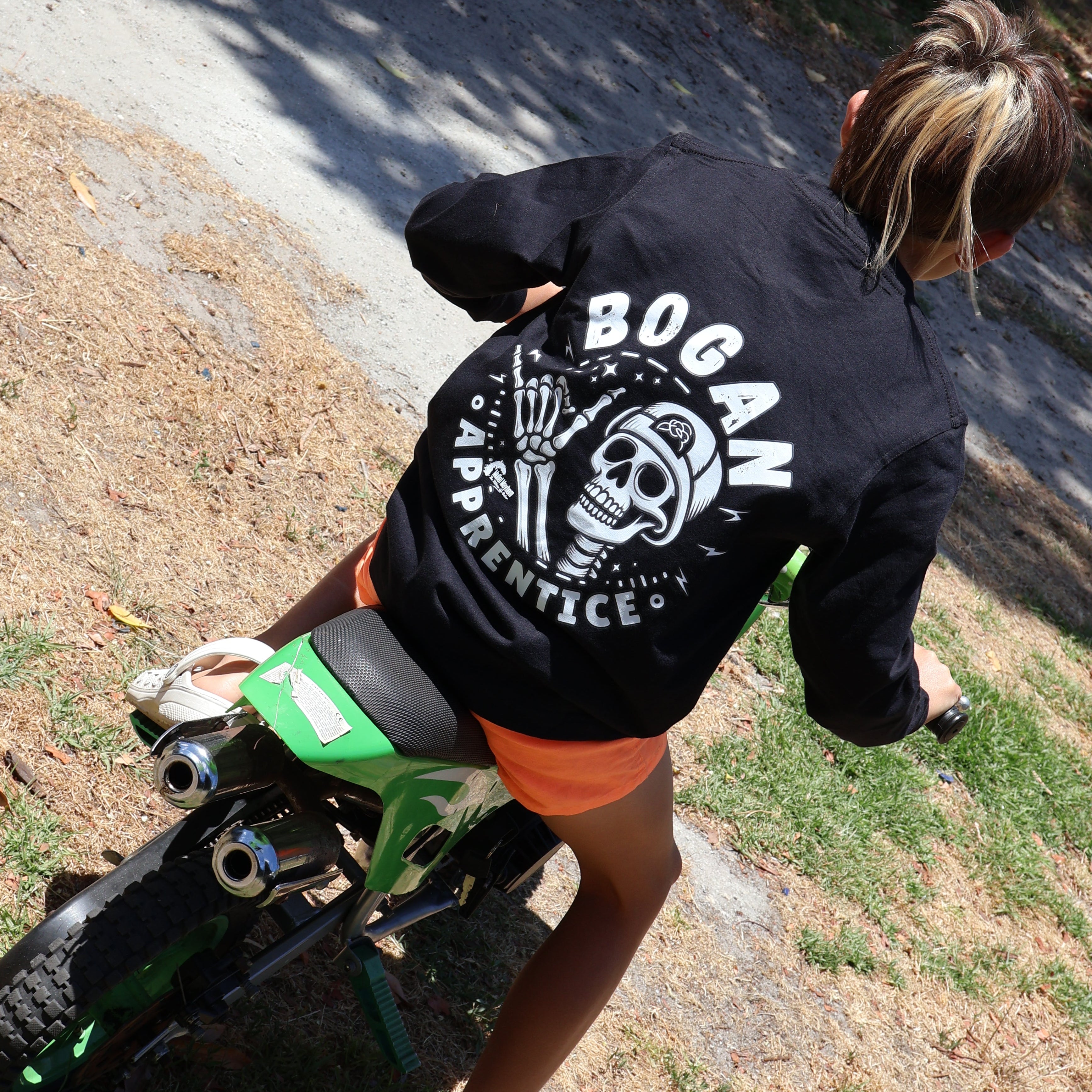 A person riding a green mini dirt bike outdoors on a driveway, wearing a black long-sleeve hoodie with a large white 'BOGAN APPLETONIC' graphic on the back and orange shorts.
