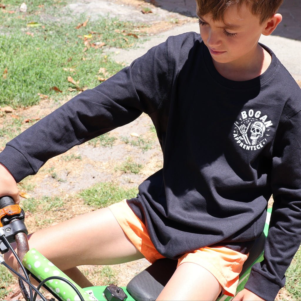 A young boy wearing a black long-sleeve crewneck shirt with a white 'BOOGA AUTOMOTIVE' skull logo on the left chest, sitting on a green tricycle outdoors on a grassy lawn.
