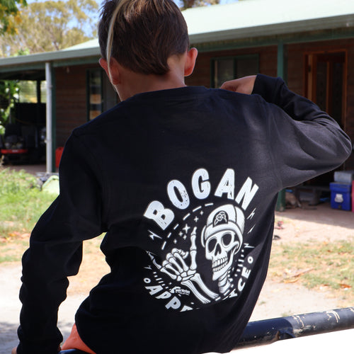 A person seen from behind wearing a black long-sleeve shirt with a large white 'BOGAN' skull graphic on the back, leaning on a vehicle outdoors in front of a house.