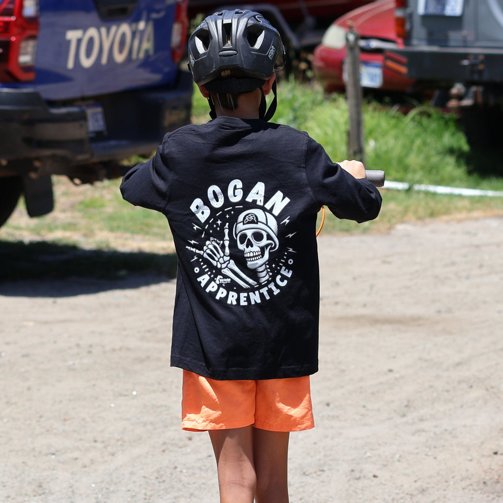 A child wearing a black Bogan Athletics hoodie, orange shorts, and a black helmet rides a scooter on a dirt path, with a blue Toyota truck and a gray SUV in the background.