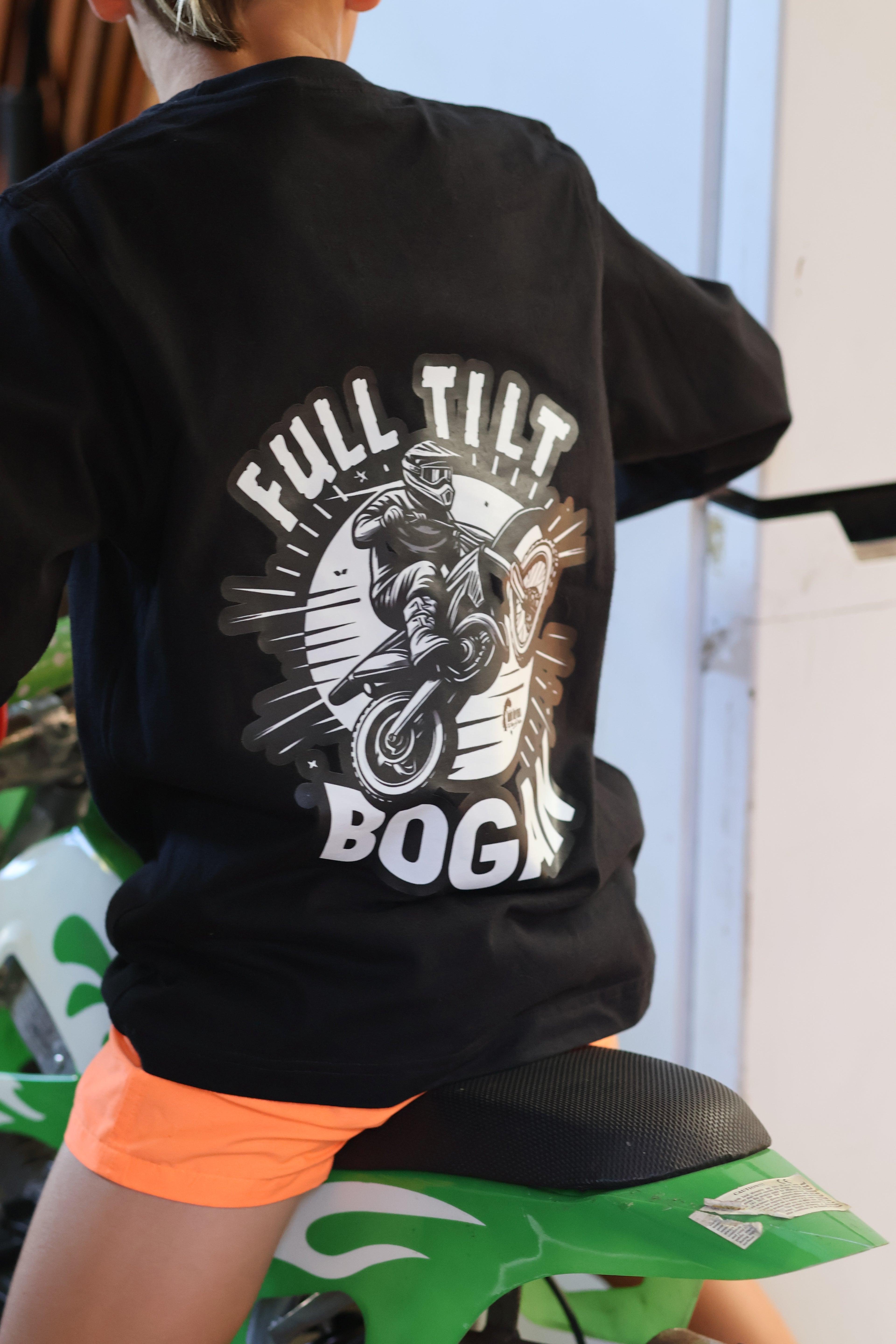 A person wearing a black graphic T-shirt with a large back print reading 'FULL TILT BOOGIE' and an illustrated motorcycle scene, seated on a green and white dirt bike in a garage-like setting.