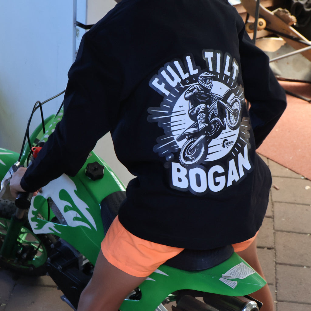 A person wearing a black long-sleeve graphic shirt with 'FULL TILT BOGAN' printed in white, sitting on a green mini dirt bike in a garage-like setting.