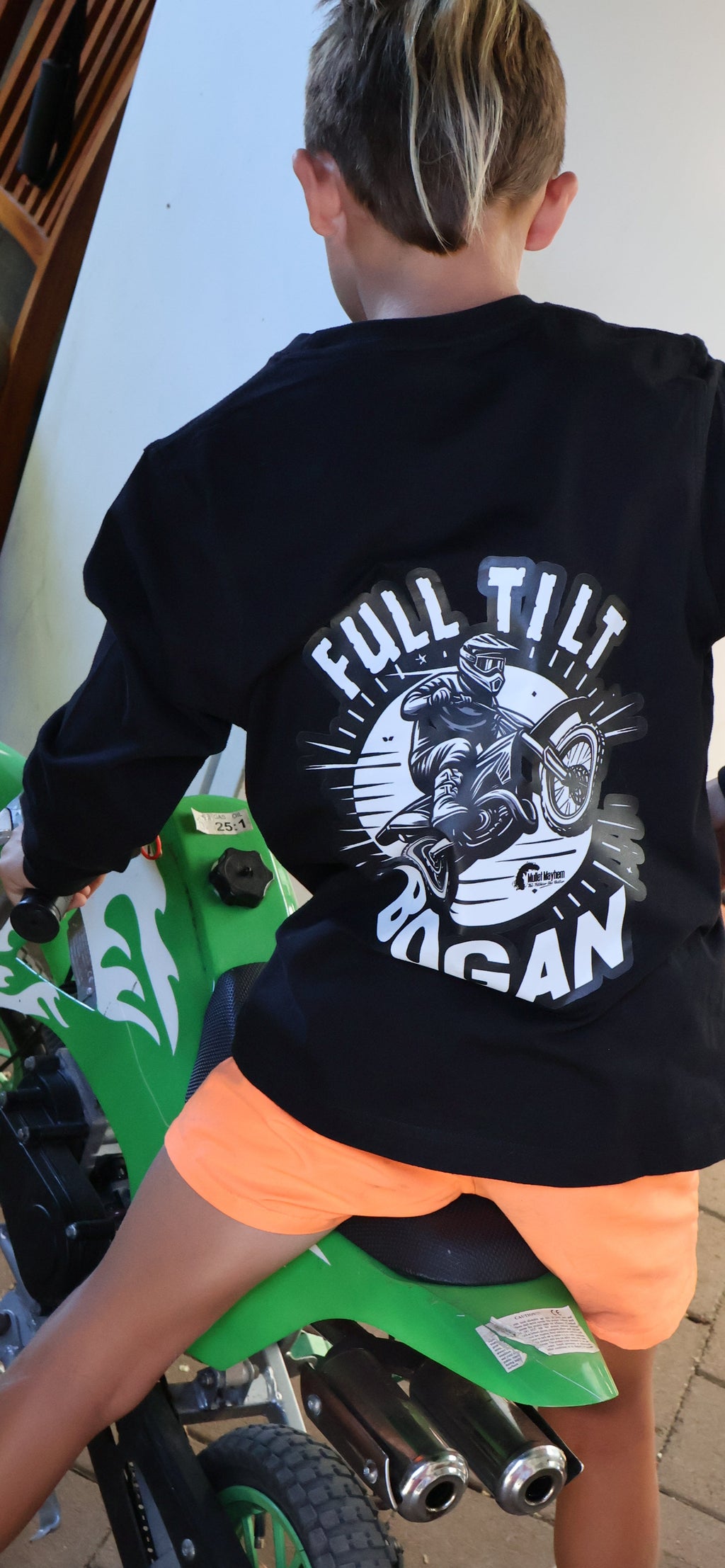 A child wearing a black long-sleeve graphic t-shirt with 'FULL TILT ROGAN' printed on the back, riding a green dirt bike in an indoor/outdoor setting.